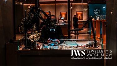 Man checking a jewellery in his stall and a person standing in front of a jewellery display behind him